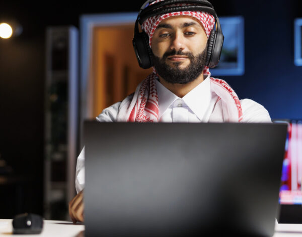 Middle Eastern man in traditional attire efficiently uses a laptop for research and communication via email. Arab guy with wireless headphones having a video call on his personal computer.