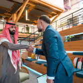 Two confident businessmen, one Middle Eastern in traditional attire and one Caucasian in a suit, shake hands in a bright, modern coworking space with wooden seating and stylish decor.