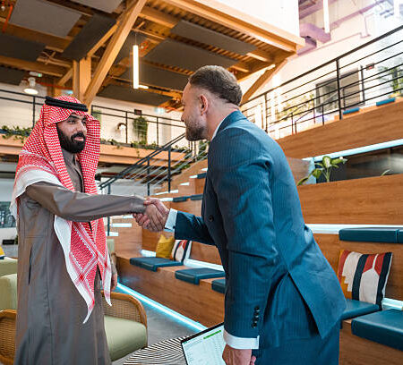 Two confident businessmen, one Middle Eastern in traditional attire and one Caucasian in a suit, shake hands in a bright, modern coworking space with wooden seating and stylish decor.