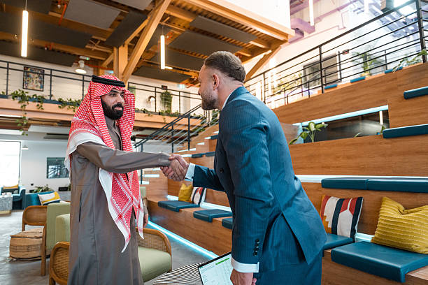 Two confident businessmen, one Middle Eastern in traditional attire and one Caucasian in a suit, shake hands in a bright, modern coworking space with wooden seating and stylish decor.