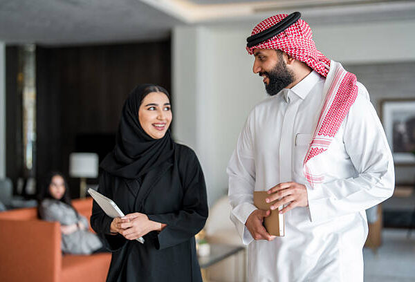 A mid adult Arabic businesswoman and businessman, dressed in traditional attire, walk confidently in a hotel lobby. The woman carries a tablet, while the man holds a smartphone, reflecting a professional atmosphere in a modern hospitality setting.