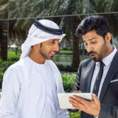 Two Arab businessmen—one wearing a traditional white kandura and the other in a formal suit—reviewing information on a digital tablet outdoors in a modern business district in Dubai, UAE. The scene conveys concepts of business collaboration, partnership, corporate communication, technology, leadership, teamwork, decision-making, and innovative entrepreneurship in the Middle East