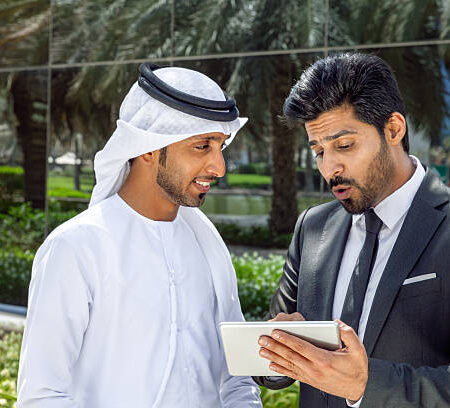 Two Arab businessmen—one wearing a traditional white kandura and the other in a formal suit—reviewing information on a digital tablet outdoors in a modern business district in Dubai, UAE. The scene conveys concepts of business collaboration, partnership, corporate communication, technology, leadership, teamwork, decision-making, and innovative entrepreneurship in the Middle East