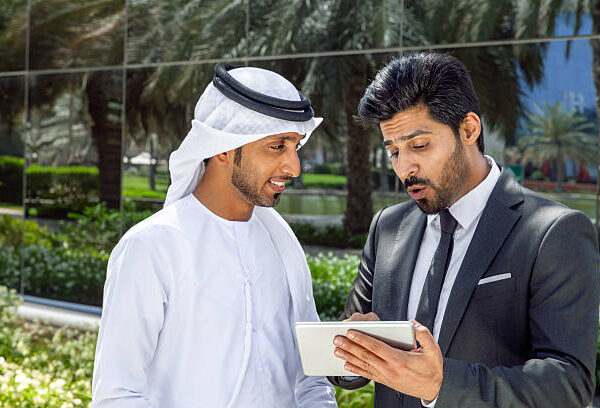 Two Arab businessmen—one wearing a traditional white kandura and the other in a formal suit—reviewing information on a digital tablet outdoors in a modern business district in Dubai, UAE. The scene conveys concepts of business collaboration, partnership, corporate communication, technology, leadership, teamwork, decision-making, and innovative entrepreneurship in the Middle East