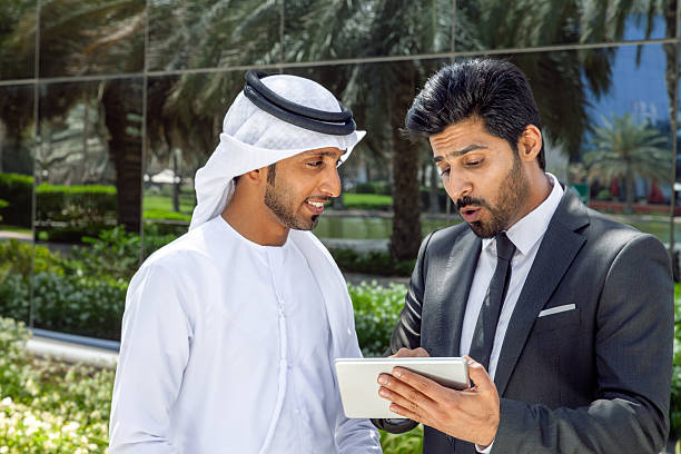 Two Arab businessmen—one wearing a traditional white kandura and the other in a formal suit—reviewing information on a digital tablet outdoors in a modern business district in Dubai, UAE. The scene conveys concepts of business collaboration, partnership, corporate communication, technology, leadership, teamwork, decision-making, and innovative entrepreneurship in the Middle East