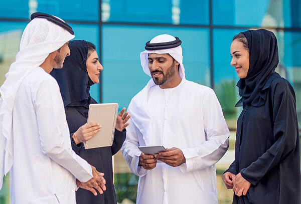 Business team of two arab businesswomen wearing abaya and two businessmen wearing kandura, traditional emirati clothes, working together in front of an office building. They are  having a conversation.